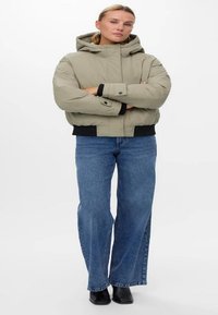 Beige dunjakke med hette, med en frontglidelås og ribbede mansjetter. Kombinert med blå vide jeans og svarte ankelstøvler.