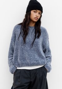 Pull en tricot bleu foncé avec une finition texturée, des poignets et un ourlet côtelés. Associé à un pantalon à rayures noir taille haute et un bonnet en tricot noir.