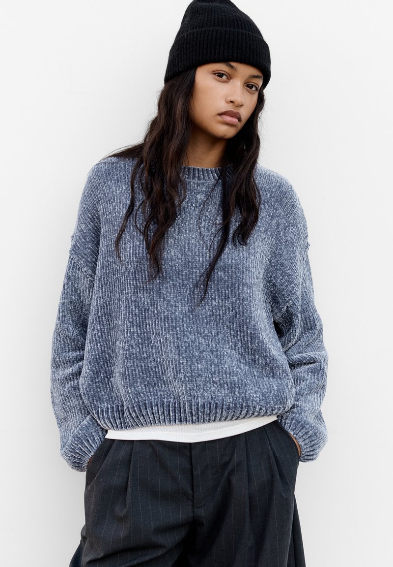 Pull en tricot bleu foncé avec une finition texturée, des poignets et un ourlet côtelés. Associé à un pantalon à rayures noir taille haute et un bonnet en tricot noir.