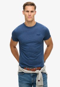 Navyblauwe korte mouwen t-shirt, ronde halslijn, gestructureerde stof, met een klein logo op de borst en een lichtgrijze trui om de taille geknoopt.