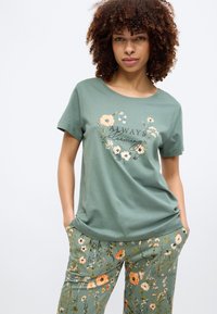 Πράσινο βαμβακερό μπλουζάκι με φλοράλ σχέδιο και το κείμενο «Πάντα Ανθίζω». Συνδυασμένο με πράσινο παντελόνι με λουλουδάτο μοτίβο για μια αρμονική εμφάνιση.