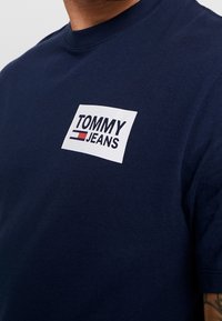 Navy blue cotton T-shirt with a round neckline and short sleeves. Features a white rectangular logo with "TOMMY JEANS" text and red/blue accents.