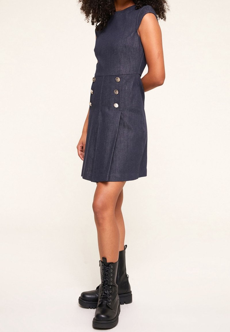 Džínové šaty v tmavě modré barvě s přiléhavým živůtkem, rukávy s krátkým střihem a dekorativními mosaznými knoflíky. Sukně má záhyby a rozšířený lem.