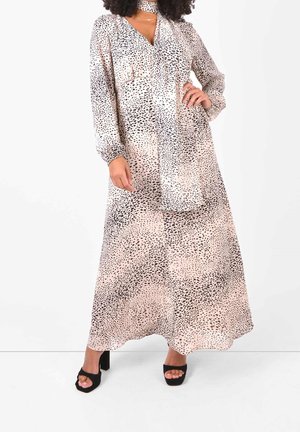 Vrouw draagt een lange, beige jurk met een zwart gestippeld patroon, lange mouwen, col met sjaal en zwarte schoenen met open teen en blokhak.