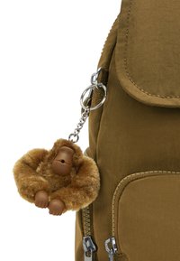Mochila caqui con un tejido texturizado, que presenta un llavero de gorila marrón y peludo sujeto por una cadena de plata, además de un bolsillo con cremallera en el lateral.