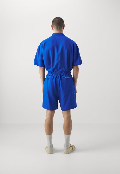 Niebieska koszula z krótkim rękawem i dopasowane szorty, wykonane z gładkiego materiału. Elastyczny pas w szortach, z małym detalem logo nad tylną kieszenią.