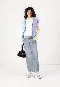 Barviti rebrasti kardigan z modrimi, sivkastimi in svetlo zelenimi deli, v kombinaciji z belim topom in dolgim jeans krilom, drži črno cvetlično torbico.