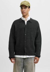 Mørkegrå strikket cardigan med et struktureret mønster, knaplukning og krave. Båret over en hvid skjorte og parret med lyse jeans.