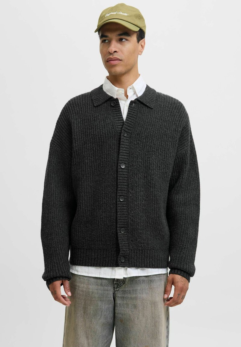 Mørkegrå strikket cardigan med et struktureret mønster, knaplukning og krave. Båret over en hvid skjorte og parret med lyse jeans.