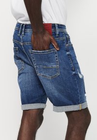 Jeans kratke hlače v srednje modrem pranju, s zavitimi robovi, petimi žepi in uničenimi detajli. Nosi se z belo srajco.