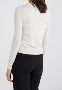 Kermanvärinen neulepaita, jossa on pitkät hihat, vartalonmyötäinen muotoilu ja ribbaustaskut. Takakuvassa näkyy sileä pinta ja lievä joustavuus.