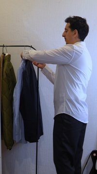 Homme en chemise blanche choisissant des chemises sur un porte-vêtements avec des vestes accrochées, devant un mur uni.