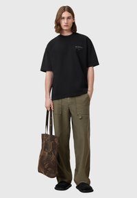 Zwarte oversized T-shirt, olijfgroene cargo broek en een bruine gestreepte schoudertas met dubbele handvaten, gedragen door een persoon met lang haar.