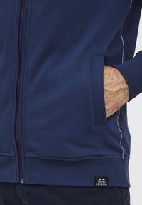 Tengerészkék, cipzáros dzseki, pamut keverékből készült, kontrasztos fehér és piros varrással, elülső zsebbel, bordázott mandzsettákkal és hem címkével.