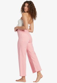 Pantaloni a gamba larga rosa chiaro realizzati in denim, con due tasche posteriori e orlo grezzo. Abbinati a un top bianco aderente.