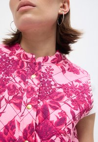 Κοντινό πλάνο ενός ατόμου που φορά ροζ εμπριμέ γιλέκο με μοβ λουλουδένια σχέδια και χρυσά τρουκ, καθώς και ασημένια σκουλαρίκια κρίκους.