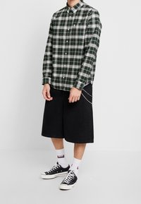 Vihreä-valkoraidallinen pitkähihainen paita, napitettu, yhdistetty mustiin polvipitkiin farkkushortseihin ja mustiin tennareihin, joissa valkoiset pohjat.