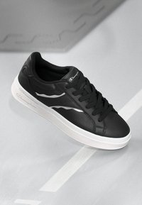 Zapatilla negra con parte superior de cuero texturizado, diseño con cordones, entresuela blanca y acentos plateados en los lados que incluyen el detalle del logo.