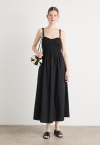 Robe maxi noire sans manches avec corsage froncé et jupe ample. Le mannequin tient un sac à main noir et crème à rayures ; elle porte des mules noires.