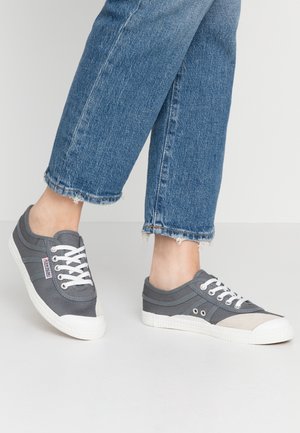 Stopy w szarych tenisówkach z płóciennego materiału z białymi podeszwami i sznurowadłami, w połączeniu ze skróconymi niebieskimi jeansami z postrzępionymi brzegami na jasnej podłodze.