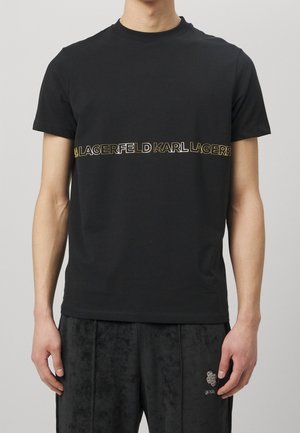 Czarna bawełniana koszulka z okrągłym dekoltem i krótkimi rękawami. Posiada złotą taśmę z logo na wysokości klatki piersiowej. Gładka faktura i dopasowany fason.