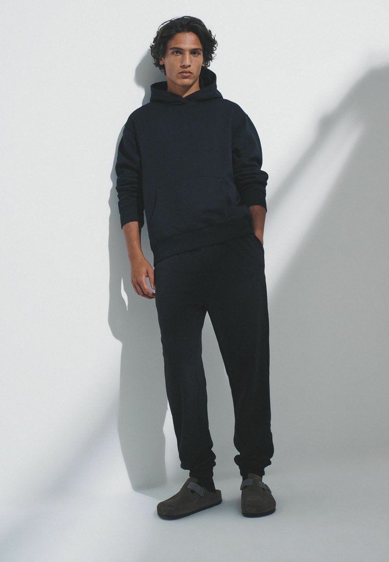 Tamsiai mėlynas džemperis su kapišonu ir priekine kišene, derinamas su tamsiai mėlynomis treniruoklio kelnėmis. Modelis dėvi rudus, lengvai užmaunamus batus su minimalistiniu dizainu.