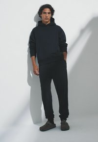 Tmavomodrá mikina s kapucňou a vreckom vpredu, doplnená zladenými teplákmi. Model má na nohách hnedé slip-on topánky s minimalistickým dizajnom.