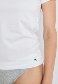Artimiausias kadras, kuriame matomas moters liemuo, vilkintis baltą marškinėlį su juodu 'CK' logotipu ir pilkas apatines kelnaites, fone - šviesi aplinka.