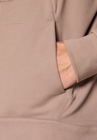 Ανοιχτό καφέ πουλόβερ με μπροστινή τσέπη και εφαρμοστά μανίκια. Μαλακό ύφασμα με ομαλή υφή, με λιτές ραφές που προσδίδουν μίνιμαλ λεπτομέρειες.