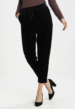 Vrouw draagt zwarte fluwelen taps toelopende broek met trekkoord in de taille, gecombineerd met zwarte schoenen met blokhakken en een glinsterende top met lange mouwen.