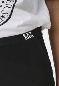 Zwarte broek met een getextureerde tailleband waarop de woorden "EXT" en "END" in witte druk staan. Ritsdetail bij de tailleband. Katoenmengselmateriaal.