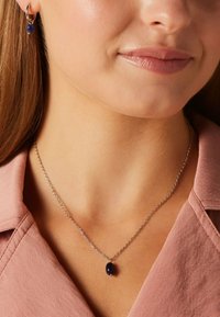 Collier en argent avec un pendentif en pierre bleu foncé en forme ovale, porté avec des boucles d'oreilles pendantes assorties. En arrière-plan, un col de chemise rose clair.