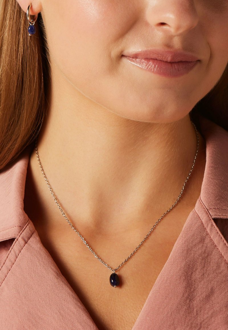 Collier en argent avec un pendentif en pierre bleu foncé en forme ovale, porté avec des boucles d'oreilles pendantes assorties. En arrière-plan, un col de chemise rose clair.