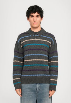 Junger Mann in einem dunkelgrauen Strickpullover mit horizontalen blauen, lila, beigen und türkisfarbenen Streifen, der in hellblauen Jeans vor einem weißen Hintergrund steht.