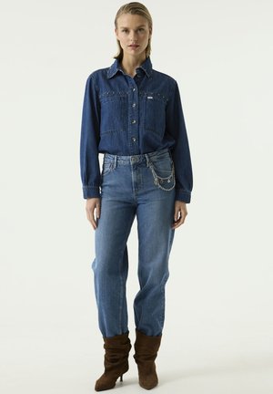 Femme debout devant un fond uni portant une chemise en jean bleue, un jean bleu avec une chaîne argentée et des bottines marron à talons.