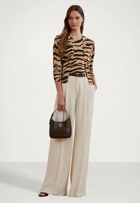 Femeie îmbrăcată cu un top și cardigan cu imprimeu de tigru maro și bej, purtând pantaloni largi crem și ținând o poșetă mică maro închis.