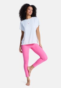 Femme aux cheveux bouclés se tient pieds nus sur une jambe, portant un haut blanc ample et des leggings rose vif sur un fond uni.