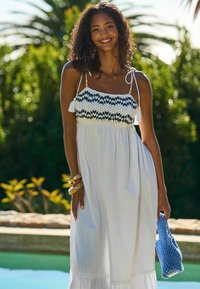 Robe d'été blanche avec un corsage tricoté à motifs noir et blanc, fines bretelles et pompons. Le mannequin tient un sac en crochet bleu.