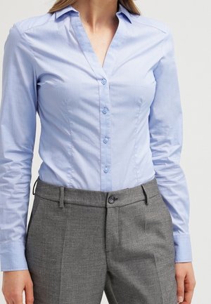 Kobieta ubrana w jasnoniebieską koszulę na guziki, włożoną do szarych spodni z wysokim stanem, na jednolitym tle.