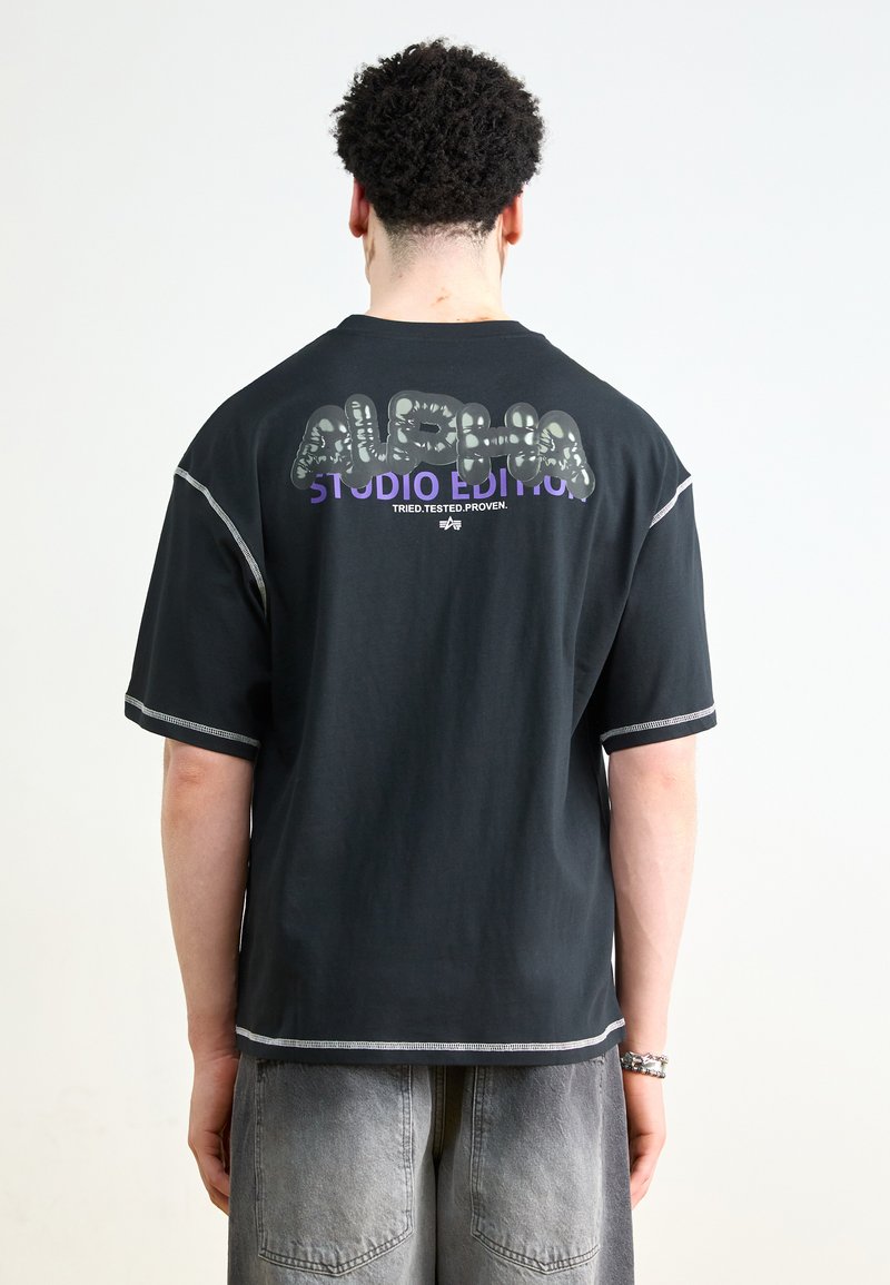 T-shirt noir à manches courtes présentant un imprimé graphique vert et violet au dos, avec des surpiqûres blanches et une coupe décontractée.
