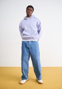 Felpa con cappuccio di colore lilla chiaro con tasca a marsupio, abbinata a jeans blu larghi. Il modello è in piedi su un pavimento giallo contro un muro bianco.