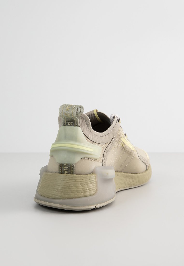 Zapatilla beige con tejido texturizado, acentos transparentes y suela acolchada. Presenta un tabón de talón con patrón y detalles en amarillo.