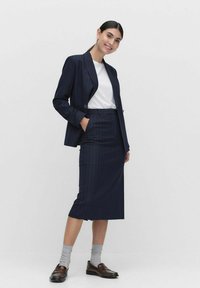 Laivansiniset raitapuvut, joissa on muotonsa mukainen bleiseri ja kynähame. Alle on puettu valkoinen t-paita. Ruskeat loafers-kengät ja harmaat sukat täydentävät asun.
