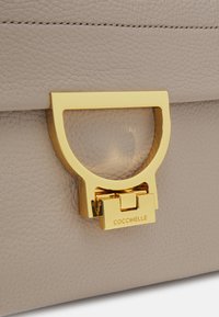 Jasnobrązowa torebka skórzana o fakturze z zapięciem z metalowym wykończeniem w kolorze złotym, wygrawerowanym napisem "COCCINELLE."