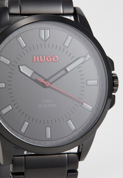 Montre-bracelet en métal noir avec un cadran gris. Les caractéristiques incluent des index horaires audacieux, des détails rouges et une résistance à l'eau jusqu'à 30 mètres.