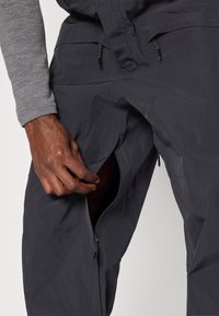 Χέρι που ξεκουμπώνει σκούρα γκρι παντελόνια με ανοιχτό φερμουάρ στη μηρή, φορώντας γκρι μακρυμάνικο μπλουζάκι.