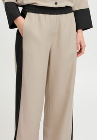 Pantalons larges en tissu beige avec des panneaux latéraux noirs. Dotés d'une taille élastique et d'une texture lisse et mate.