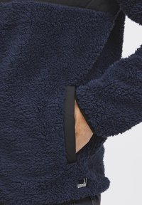Navy fleece pullover with a plush texture, featuring a black panel and pockets. Includes branding tag on the lower side.