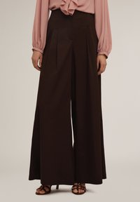 Breda byxor i mörkbrunt tyg med framfållor, ihop med en ljusrosa blus. Slät textur, minimalistisk design, avslappnad passform.