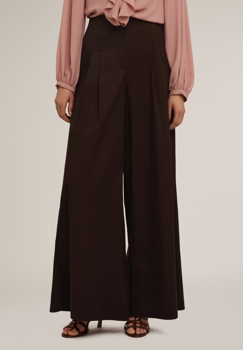 Spodnie o szerokich nogawkach w ciemnobrązowym materiale z plisami z przodu, zestawione z jasnoróżową bluzką. Gładka faktura, minimalistyczny design, luźny krój.